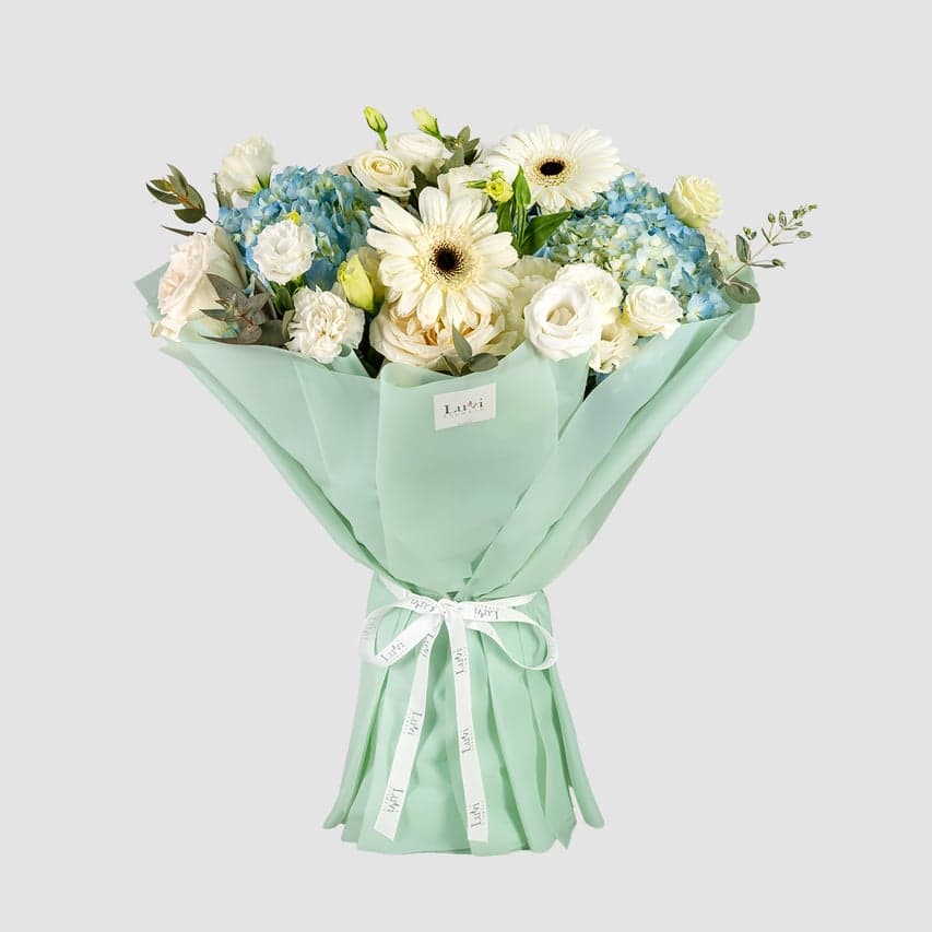 باقة توهج الندى تضم الجربيرا البيضاء، زهور الهيدرنجيا الزرقاء، الليسيانثس الأبيض، ورود أوهارا البيضاء، القرنفل الأبيض، والكافور