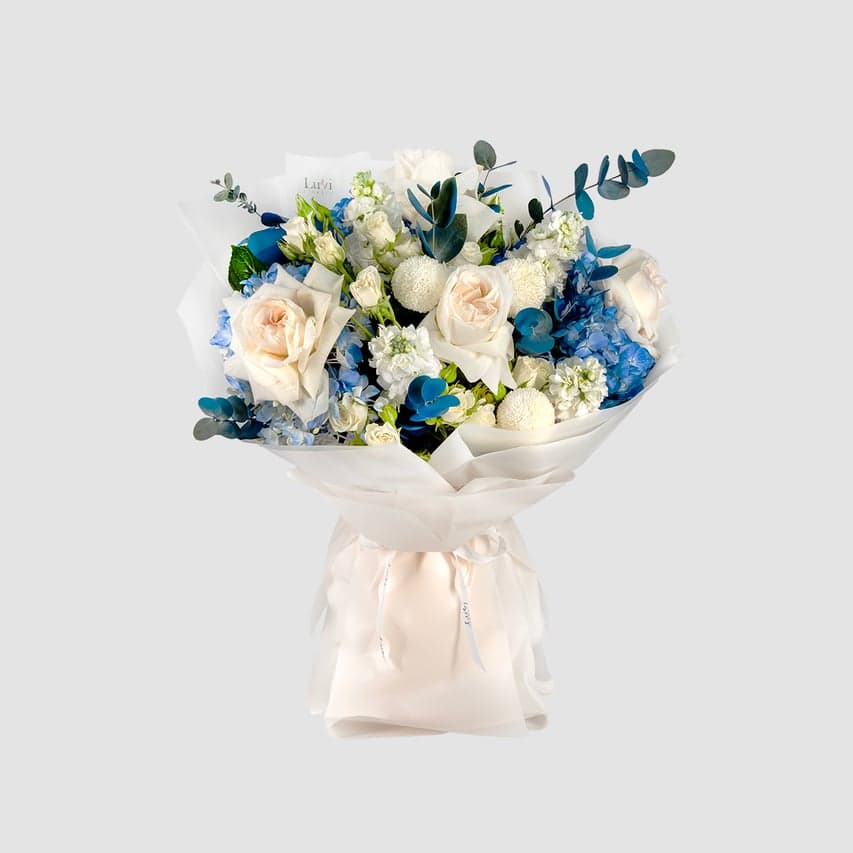 Peaceful Grace bouquet featuring blue sprayed eucalyptus, white Ohara roses, blue hydrangeas, white spray roses, white ping pong chrysanthemums, and white matthiola	