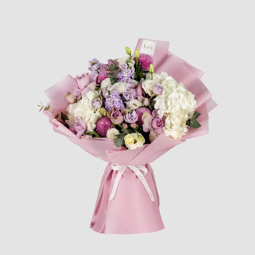 باقة سحر الاحتفال تضم زهور الهيدرنجيا البيضاء، أقحوان البينغ بونغ الأرجواني، الورود الأرجوانية، الليسيانثس الأبيض، ورود الرش الأرجوانية، والماثيولا الأرجوانية